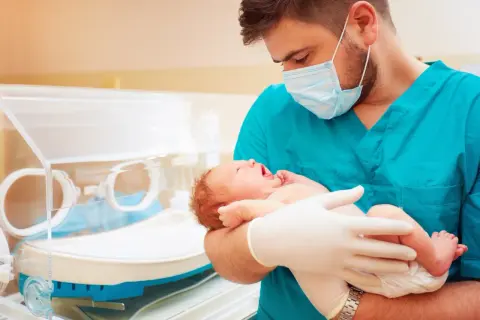Lekarz w maseczce i rękawiczkach trzyma noworodka. To obrazek o tematyce neonatologia co to, pokazujący troskę o najmniejszych.