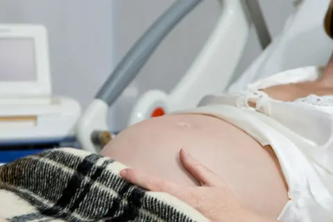 Kobieta w szpitalnym łóżku, z ręką na brzuchu, czeka na poród. Obok monitor KTG, który pomaga ocenić stan dziecka i może sugerować, jak przyspieszyć poród.