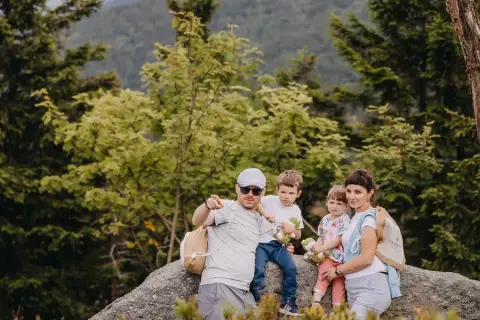 Rodzina na skale w lesie, świętująca 30 urodziny. Tata wskazuje palcem, mama tuli córkę, a synowie trzymają pluszaki.