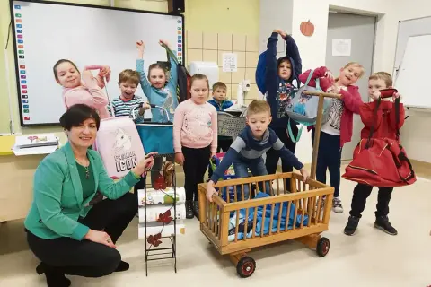 Dzień bez plecaka pomysły: dzieci z panią prezentują kreatywne sposoby przenoszenia rzeczy, od wózeczka po wielką czerwoną torbę.