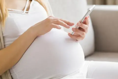 Kobieta w 24. tygodniu ciąży przegląda coś na telefonie, siedząc na kanapie.