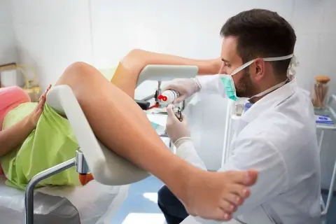 Lekarz bada pacjentkę, która leży na fotelu ginekologicznym. Widoczne są nogi pacjentki i narzędzia medyczne. Obraz pokazuje, jak wygląda zszyte krocze po porodzie.