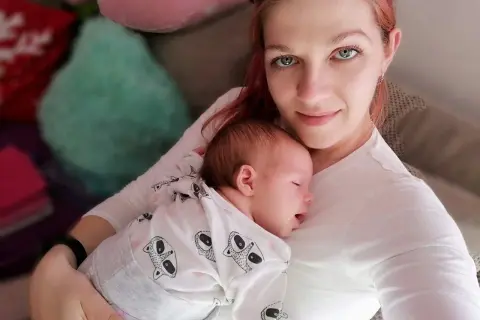 Mama tuli otula swoje śpiące dziecko, czułe kangurowanie dziecka w białym pajacyku z motywem zwierzątek.