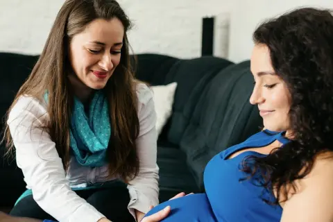 Doula wspiera przyszłą mamę. Kobieta w niebieskiej sukience czule dotyka brzucha, a obok siedzi doula, uśmiechając się ciepło.