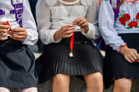Dzieci w szkolnych strojach trzymają dzwoneczki na czerwonej wstążce. Czy to symbol końca roku i nadzieja na średnią na czerwony pasek w 4 klasie?