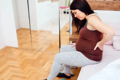 Kobieta w 27 tygodniu ciąży siedzi na łóżku, masując bolący odcinek lędźwiowy.
