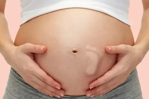 Brzuszek w 29 tygodniu ciąży, z widocznym odciskiem stópki dziecka. Ręce delikatnie obejmują brzuch.