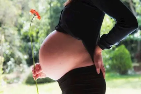 Brzuch kobiety w zaawansowanej ciąży, prawdopodobnie w 3 trymestrze. Trzyma ona pomarańczowy kwiat.
