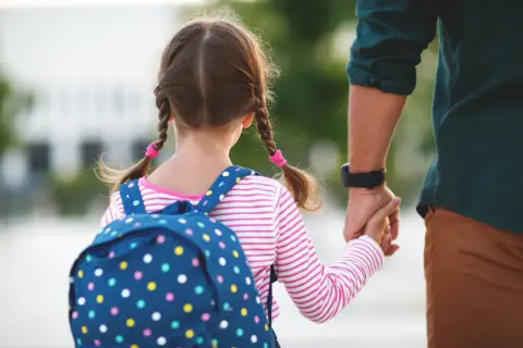 Dziewczynka z plecakiem i warkoczykami trzyma tatę za rękę. To piękny dzień, idealny na urlop okolicznościowy.