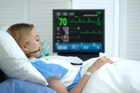 Kobieta w masce tlenowej leży w szpitalnym łóżku, monitor pokazuje parametry życiowe. Jej stan budzi niepokój, być może to rzucawka.