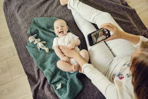 Uśmiechnięte niemowlę do kiedy będzie tak radosne? Mama robi mu zdjęcie telefonem, leży na kocyku z zabawkami.