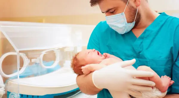 Lekarz w maseczce i rękawiczkach trzyma noworodka. To obrazek o tematyce neonatologia co to, pokazujący troskę o najmniejszych.