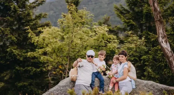 Rodzina na skale w lesie, świętująca 30 urodziny. Tata wskazuje palcem, mama tuli córkę, a synowie trzymają pluszaki.