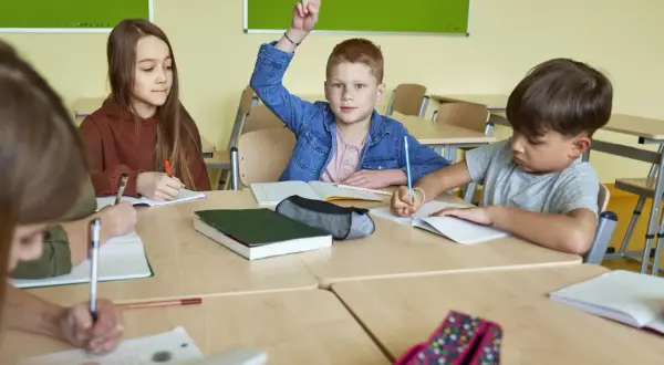 Chłopiec z podniesioną ręką, gotów odpowiedzieć na pytanie: "Od ilu procent jest 3?". Dzieci piszą w zeszytach.
