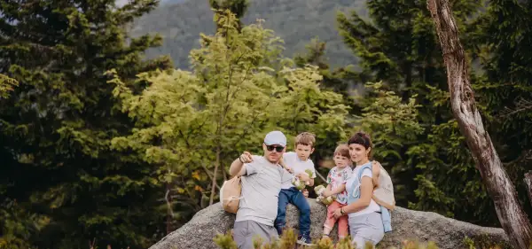 Rodzina na skale w lesie, świętująca 30 urodziny. Tata wskazuje palcem, mama tuli córkę, a synowie trzymają pluszaki.