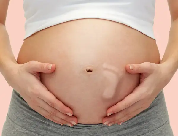 Brzuszek w 29 tygodniu ciąży, z widocznym odciskiem stópki dziecka. Ręce delikatnie obejmują brzuch.