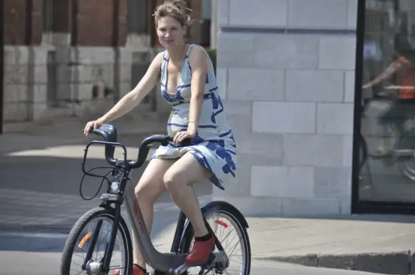 Uśmiechnięta ciężarna kobieta w letniej sukience jeździ na rowerze miejskim, pokazując, że **czy w ciąży można jeździć na rowerze** jest możliwe.
