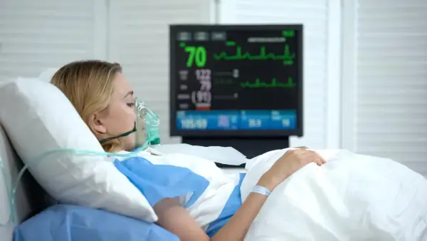 Kobieta w masce tlenowej leży w szpitalnym łóżku, monitor pokazuje parametry życiowe. Jej stan budzi niepokój, być może to rzucawka.