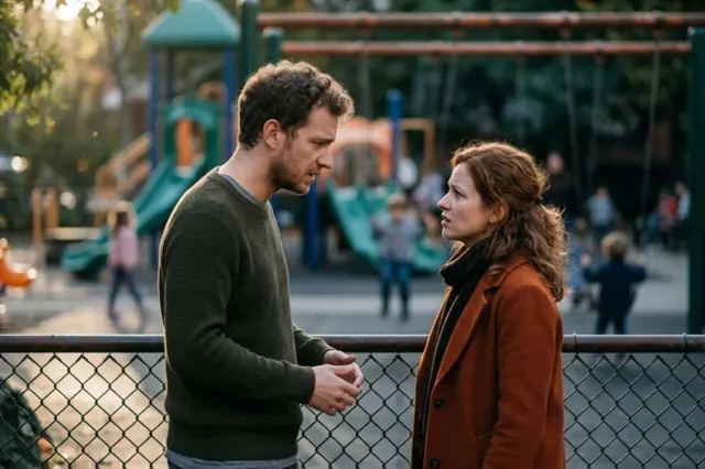 Para stoi przy płocie placu zabaw, w tle bawią się dzieci. Mężczyzna i kobieta rozmawiają, w ich oczach widać ból i smutek, sugerując zdrada.